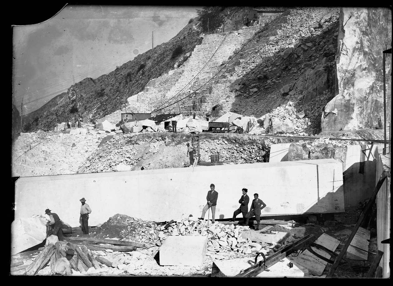 Drei Männer beobachten die Arbeit mehrerer Bagger in den Apuanischen Alpen. Hinter ihnen befindet sich ein riesiger Marmorblock, der bereits quadratisch zugeschnitten und bereit ist, ins Tal transportiert zu werden von A. Corsini