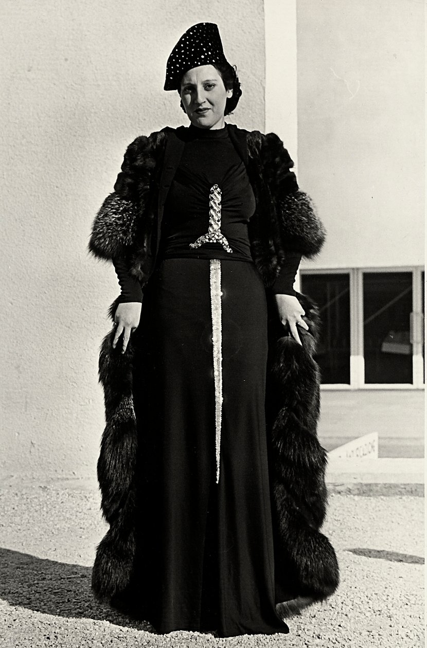 Modell im Abendkleid des italienischen Modehauses "Dragoni". Die Vorderseite des Artikels hat eine auffällige Paillettenverzierung in Form eines Schwertes. Ein Hut und ein Pelzmantel vervollständigen das Ensemble. von Luis Bogino