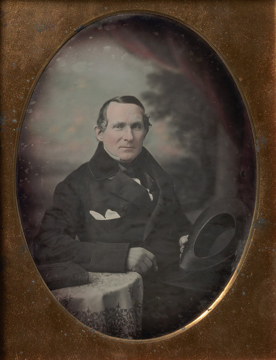 Mann mit Mantel und Handschuhen, der Hut hält, vor gemaltem Outdoor-Hintergrund sitzend von Samuel Broadbent Jr..