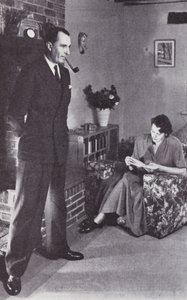 Frau liest, während der Mann sich am Feuer den Rücken wärmt, ca. 1950 von English Photographer