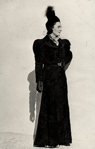 Modell präsentiert einen Artikel des italienischen Modehauses "Cicogna" bestehend aus einem Breitschwanzmantel, der an der Taille mit einem Gürtel mit juwelenbesetzter Schnalle zusammengehalten wird, ergänzt durch einen kleinen Federhut mit Schleier. von Luis Bogino