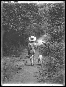 William Gray Hassler mit Strohhut und Latzhose, trägt eine Angelrute und einen Korb, geht einen Weg im Wald entlang mit einem kleinen Hund (Bessie), 20. August 1911
