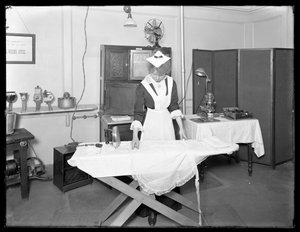 Junge Frau in einem Dienstmädchenkostüm demonstriert die Verwendung eines elektrischen Bügeleisens, ca. 1913-14
