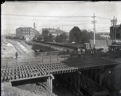 Horizontales Schwarz-Weiß-Foto einer Holzbrücke und Straßenbahnschienen über teilweise gebaute Eisenbahnschienen. Eine Straßenbahn mit der Aufschrift Chouteau 2137 beginnt rechts die Brücke zu überqueren. Im Hintergrund Fabrikgebäude mit der Aufschrift Sta von Charles Clement Holt