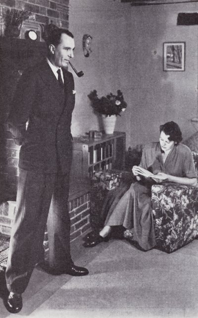 Frau liest, während der Mann sich am Feuer den Rücken wärmt, ca. 1950 von English Photographer