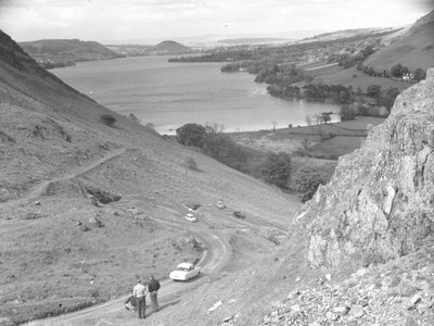 Ein Blick hinunter auf eine Hügelseite mit Straße, Autos und Menschen, See im Hintergrund von Joseph Hardman