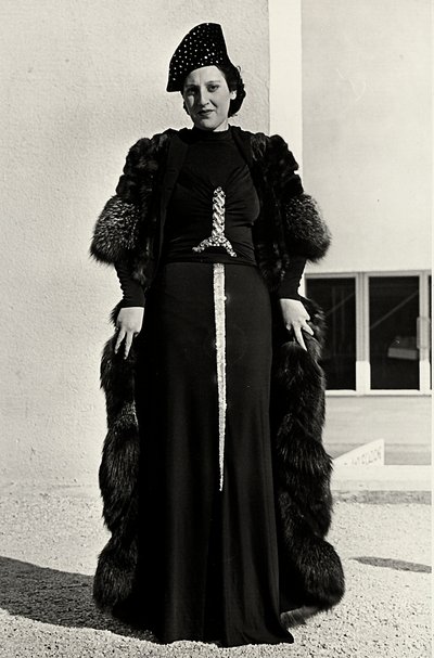 Modell im Abendkleid des italienischen Modehauses "Dragoni". Die Vorderseite des Artikels hat eine auffällige Paillettenverzierung in Form eines Schwertes. Ein Hut und ein Pelzmantel vervollständigen das Ensemble. von Luis Bogino