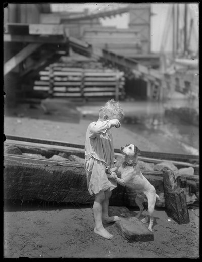 Barfüßiger Junge, möglicherweise William Gray Hassler, spielt mit Hund am Ufer eines Flusses oder Sees, New York, ca. 1910 von William Davis Hassler