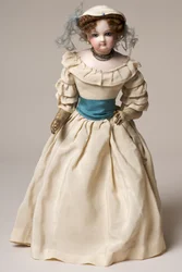 Biskuitmodepuppe mit schwenkbarem Kopf, trägt ein langes cremefarbenes Kleid und einen Hut