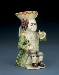 Toby-Krug eines Zechers, sein Hut mit braun-gelb gebogtem Rand, sein eingeritztes braunes Haar zum Zopf gebunden und mit einer weißen Rosette, sitzend mit einem Balusterkrug auf dem Knie, ca. 1770