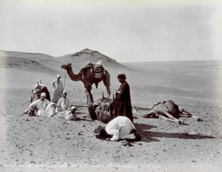 Ägyptische Kamelreiter während einer Pause in der Wüste. Im Vordergrund scheinen zwei Mitglieder der kleinen Gruppe zu beten