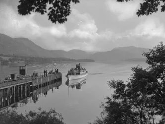 Ein Boot mit Menschen auf einem See nähert sich einem Steg, Hügel im Hintergrund
