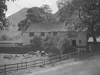 Ein Gebäude vor dem Kälber weiden, rechts stehen zwei Frauen und unterhalten sich an einer Wand, im Hintergrund Hügel