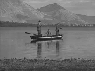 Drei Männer in einem Ruderboot auf einem See beim Angeln, ein Mann sitzt, Hügel im Hintergrund