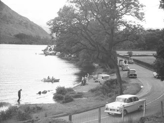 Blick auf das Seeufer mit Menschen, die stehen, gehen, waten und Boot fahren, Straße verläuft rechts im Bild mit geparkten Autos