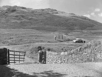 Blick auf die Straße über den Hard Knott Pass, zwei Personen stehen in der Nähe eines Straßenschilds und ein geparktes Auto auf dem Grasstreifen