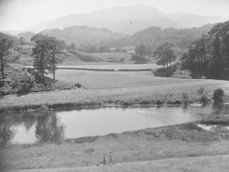 Frauen schieben Baby im Kinderwagen am Fluss entlang durch eine Wiese mit Menschen, die im Fluss schwimmen, und Bauer, der Traktor auf Feldern im Hintergrund arbeitet