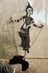 Ein Puppenspieler führt vor einer Gaze eine erzählende Schattenfigur für ein Schattenspiel. C…
