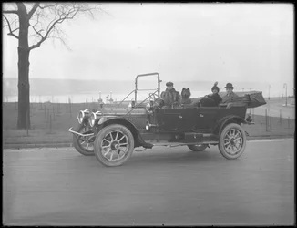 Drei nicht identifizierte Personen und zwei Chow-Hunde fahren in einem Cabriolet entlang eines Flusses oder Sees, ca. 1912