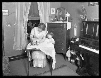 Unidentifizierte Frau demonstriert die Verwendung eines elektrischen Bügeleisens, beobachtet von kleinem Mädchen, ca. 1913-14